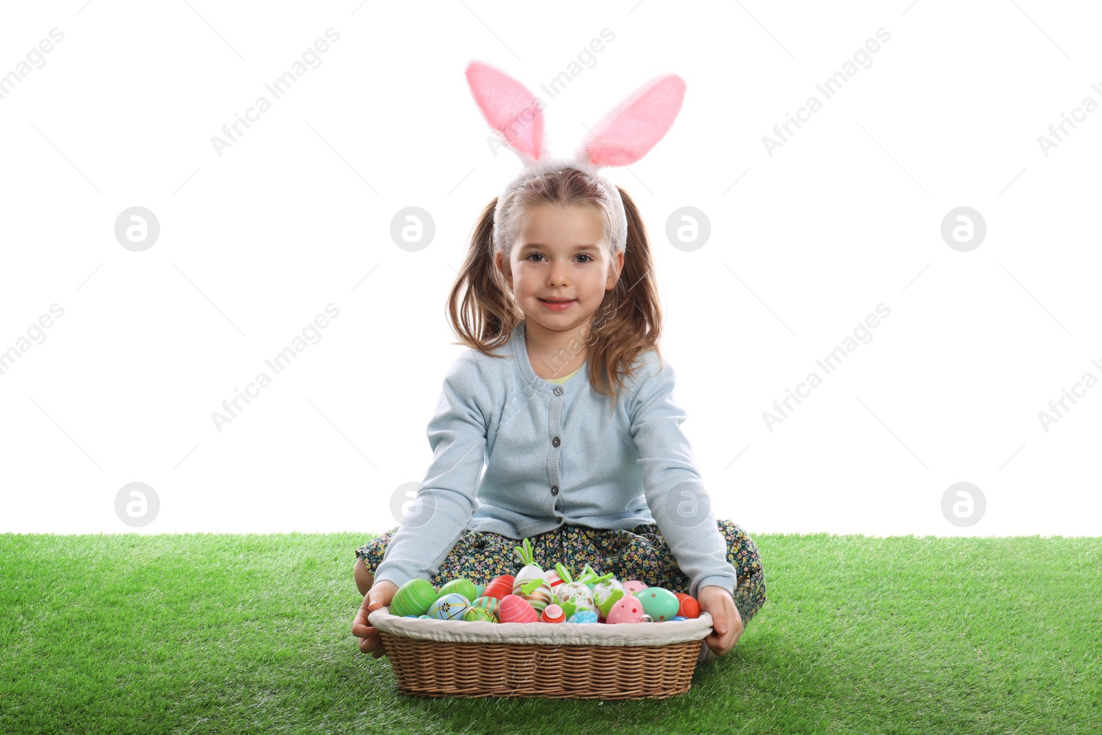 Photo of Adorable little girl with bunny ears and wicker basket full of Easter eggs on green grass against white background