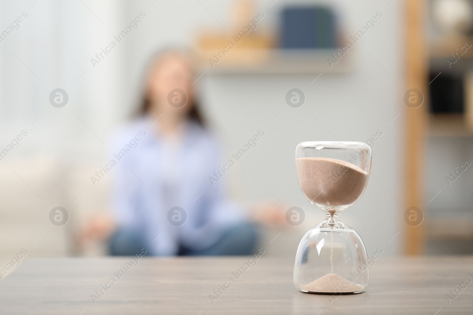 Photo of Hourglass with flowing sand on desk. Woman meditating indoors, selective focus