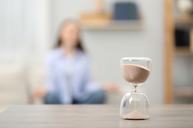 Photo of Hourglass with flowing sand on desk. Woman meditating indoors, selective focus