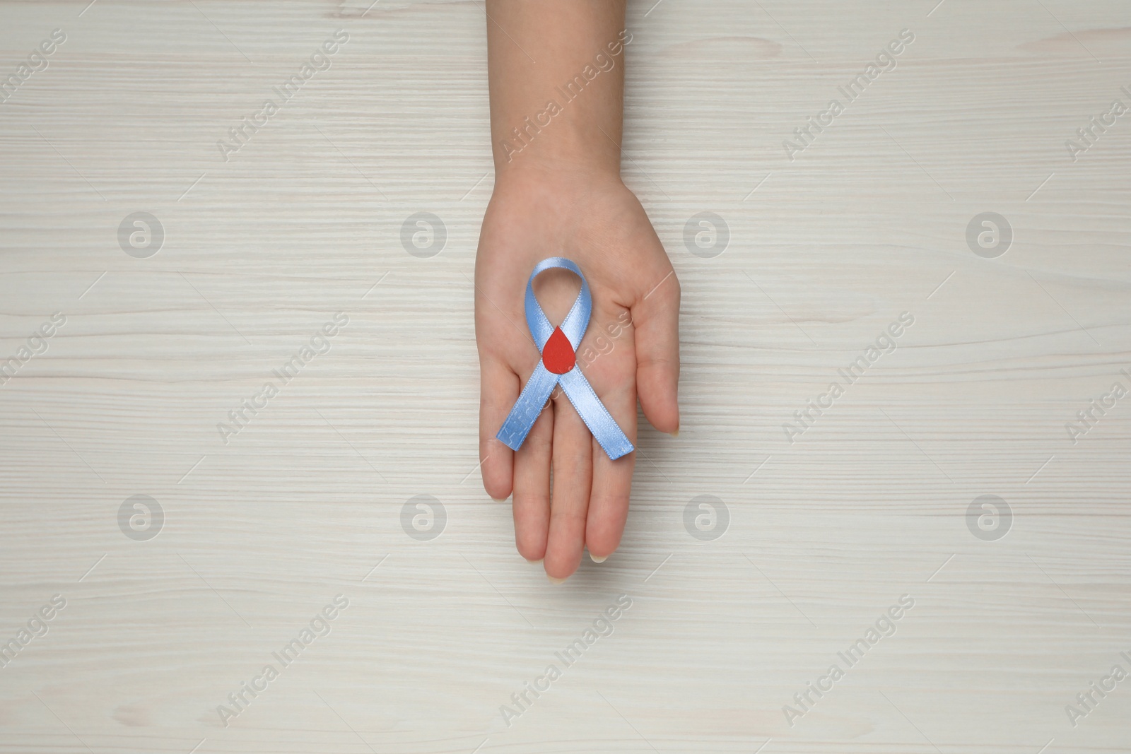 Photo of Woman holding light blue ribbon with paper blood drop at wooden table, top view. Diabetes awareness