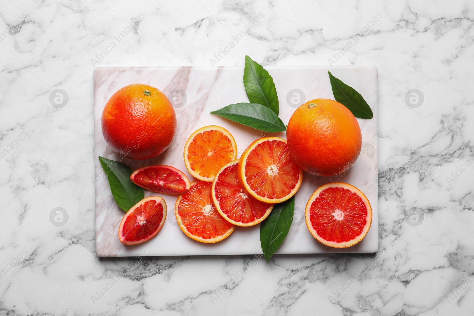 Photo of Whole and cut red oranges with green leaves on white marble table, top view