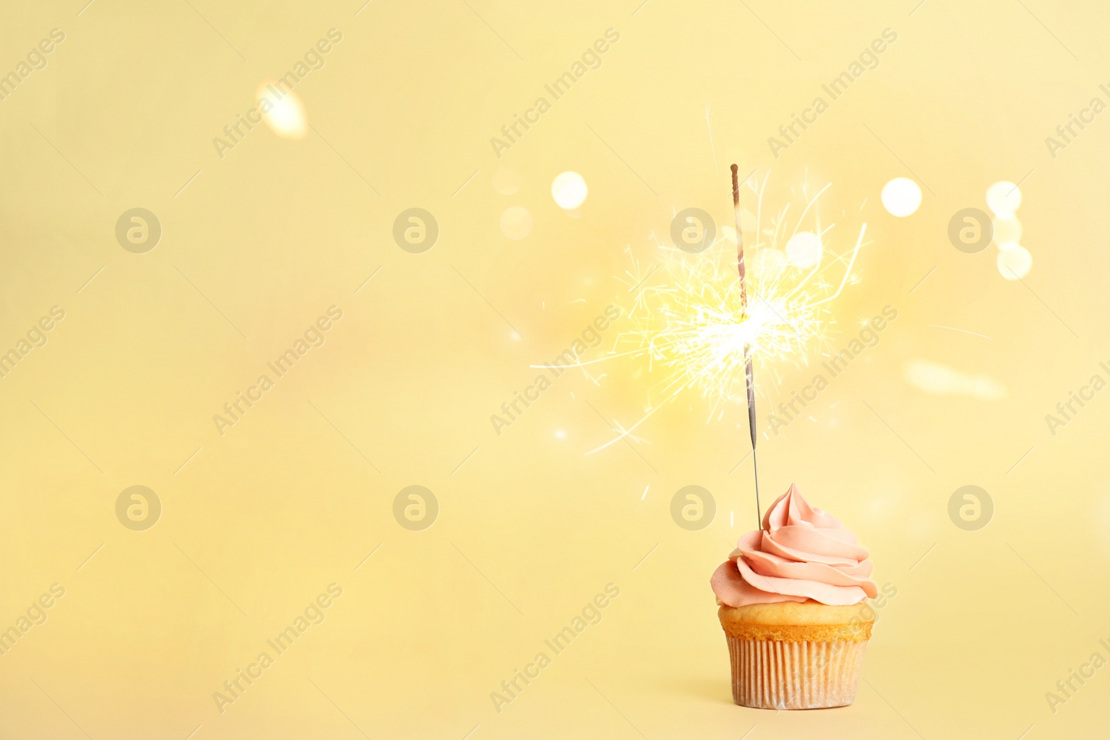 Image of Birthday cupcake with sparkler on light yellow background. Space for text