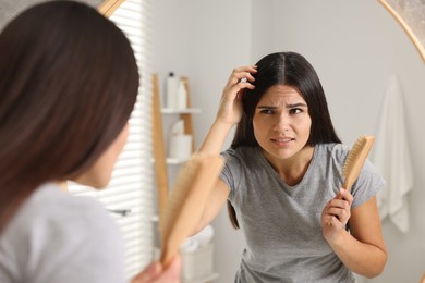 Photo of Emotional woman with brush examining her hair and scalp near mirror in bathroom. Dandruff problem