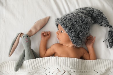 Photo of Adorable newborn baby in knitted hat with toy bunny on bed, top view