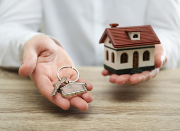 Photo of Real estate agent holding house model and key at wooden table, closeup