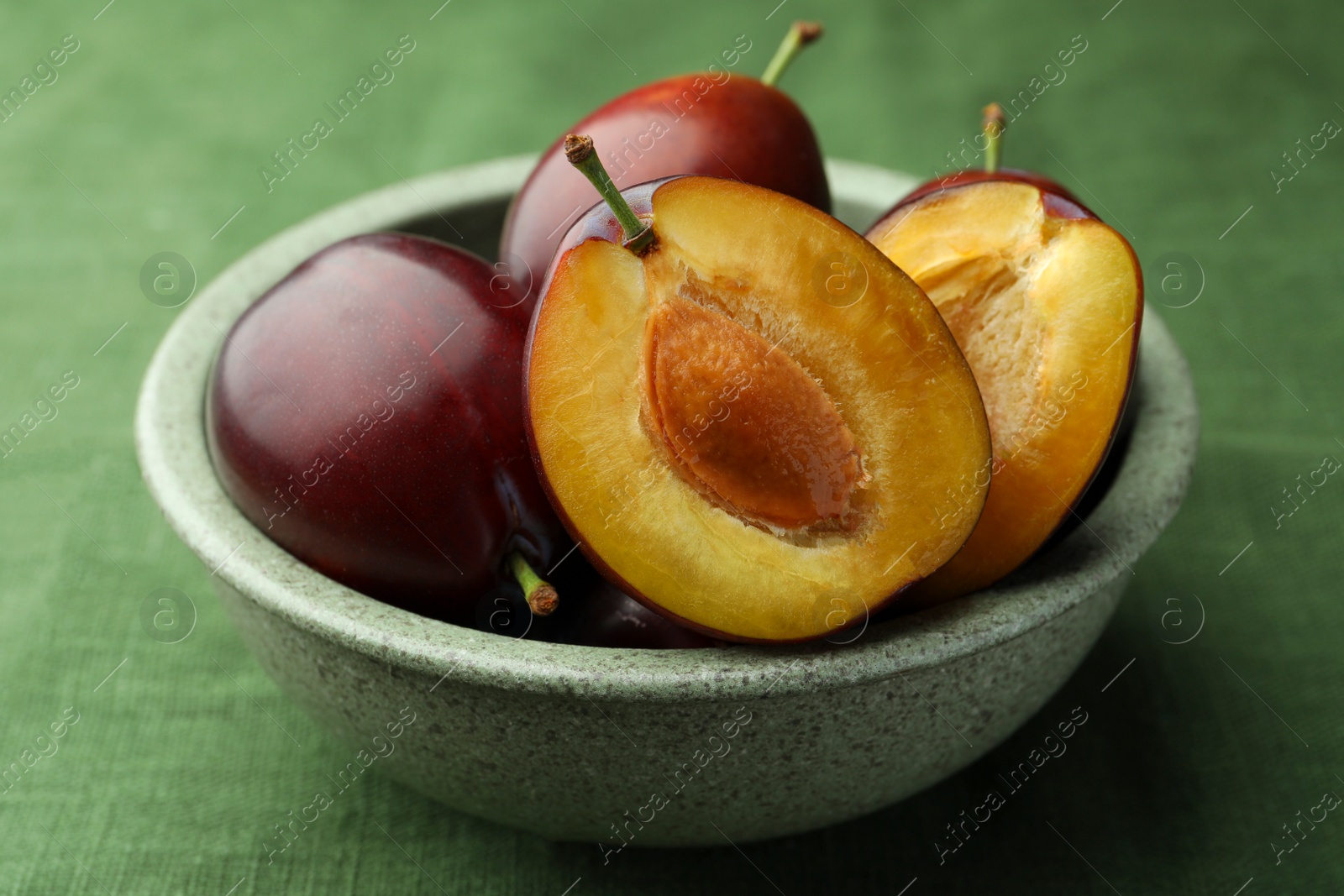 Photo of Many tasty ripe plums in bowl on green fabric, closeup