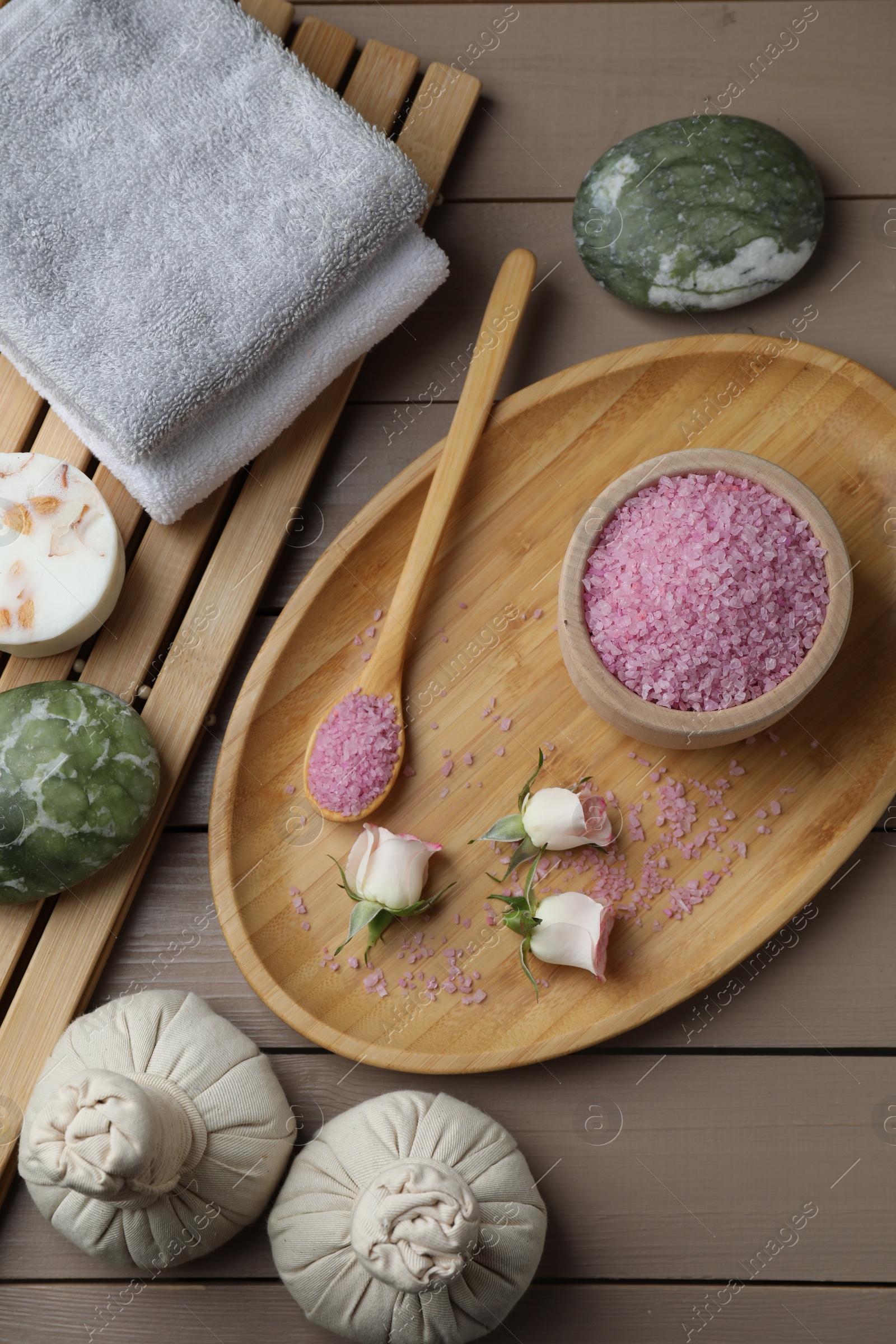 Photo of Bowl of pink sea salt, roses, spa stones, herbal massage bags and towels on wooden table, flat lay