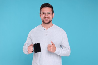 Photo of Portrait of happy man holding black mug and showing thumb up on light blue background