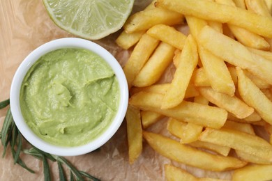 Delicious french fries, avocado dip, lime and rosemary on parchment, top view