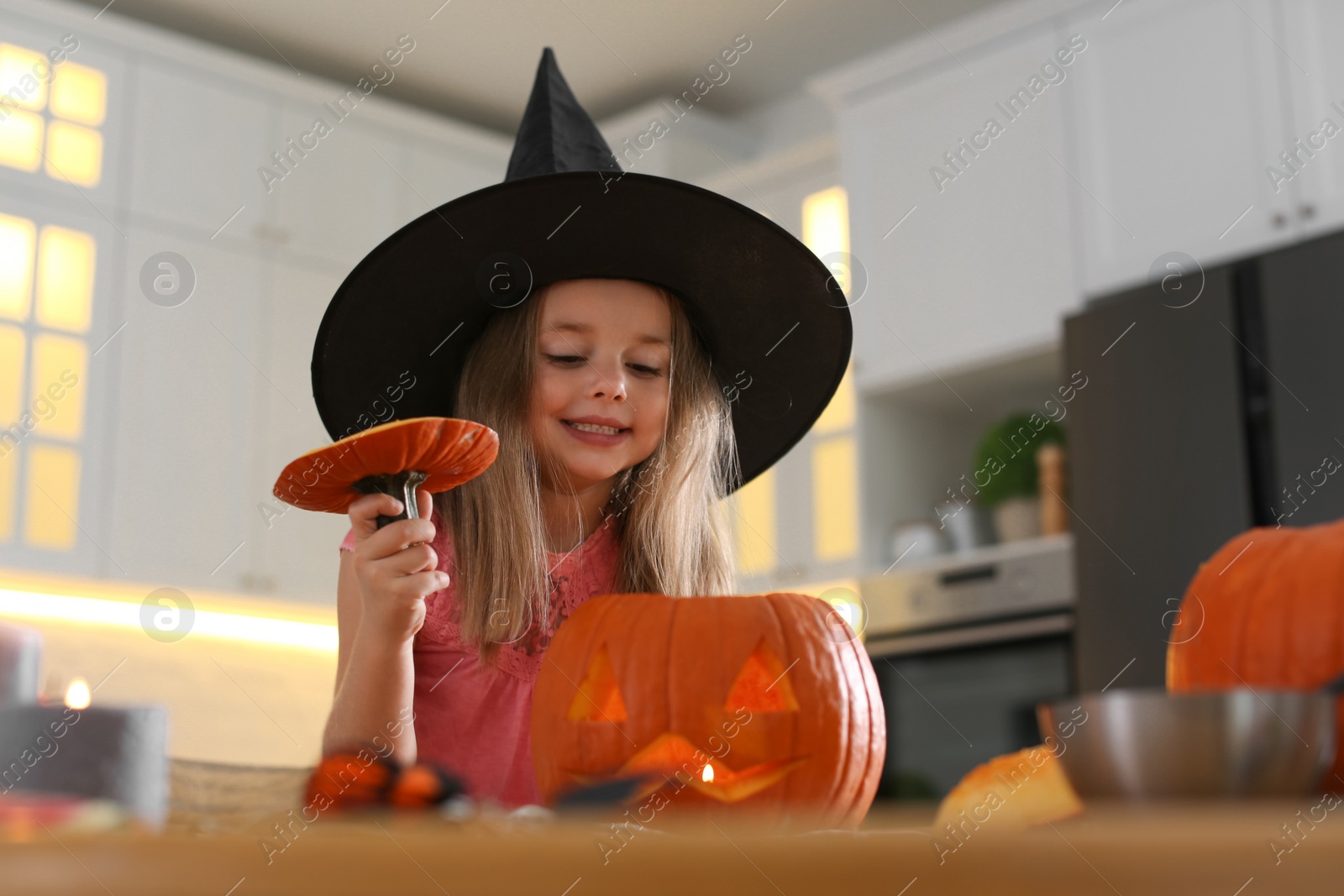 Photo of Little girl with pumpkin jack o'lantern at table in kitchen. Halloween celebration