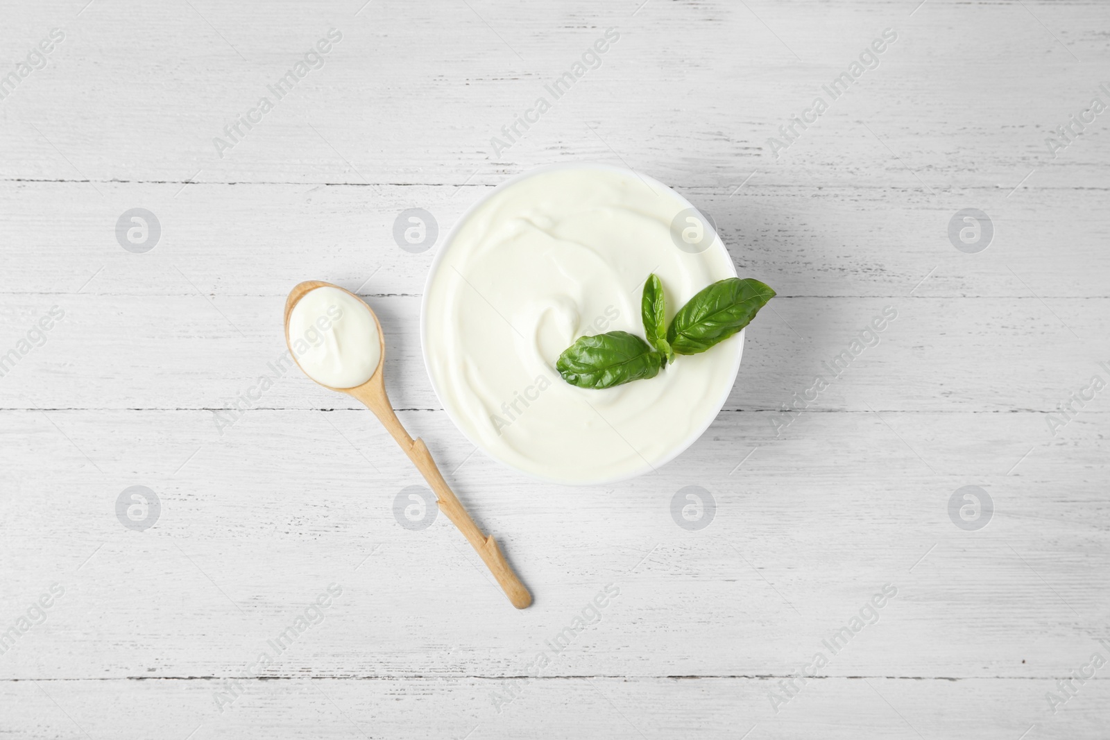 Photo of Bowl of fresh sour cream with basil and spoon on white wooden table, flat lay