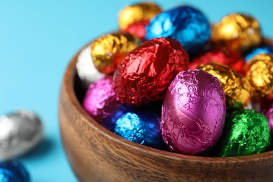 Wooden bowl with chocolate eggs wrapped in colorful foil on light blue background, closeup