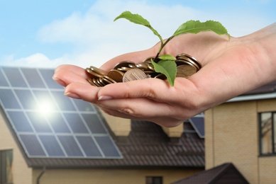 Image of Woman holding coins and green sprout against house with installed solar panels on roof, closeup. Economic benefits of renewable energy