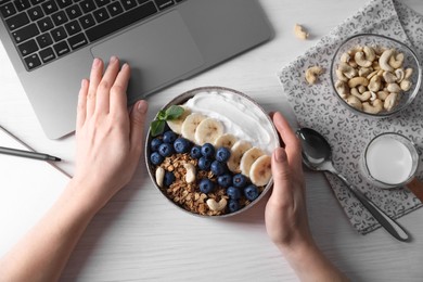 Woman with bowl of tasty granola working with laptop at white wooden table, top view