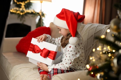Cute little girl opening gift box on sofa in room decorated for Christmas