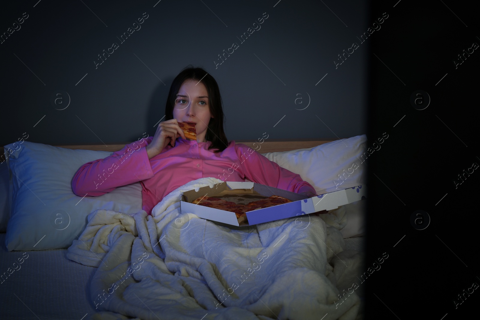 Photo of Young woman eating pizza while watching TV on bed at night