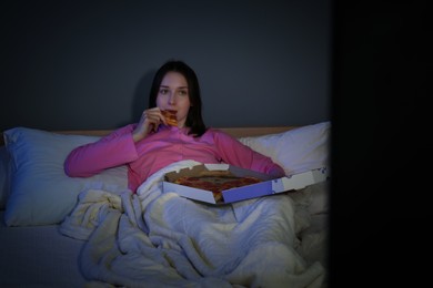 Photo of Young woman eating pizza while watching TV on bed at night