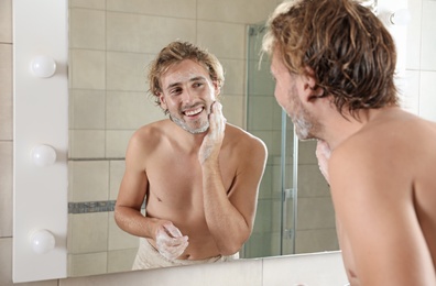 Photo of Young man washing face with soap near mirror in bathroom