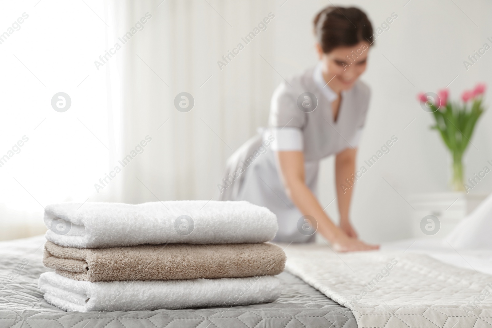 Photo of Young maid making bed in hotel room, focus on stack of towels