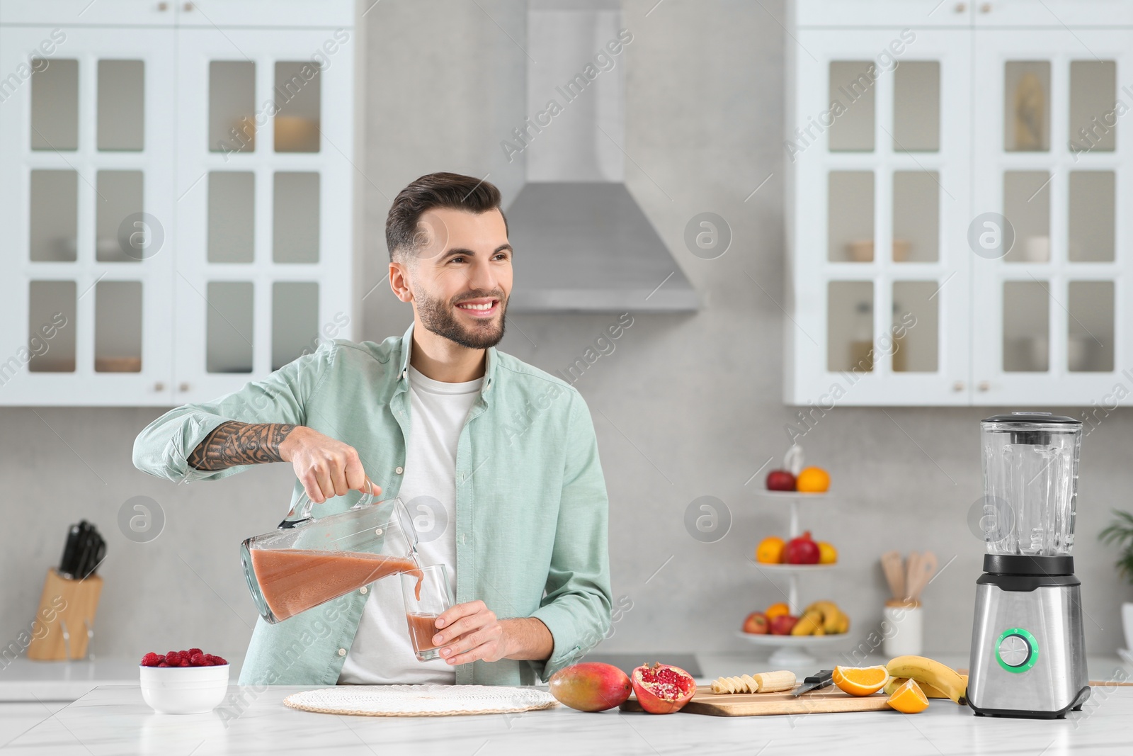 Photo of Handsome man pouring tasty smoothie into glass at white marble table in kitchen. Space for text