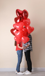 Photo of Romantic couple hiding behind heart shaped balloons near beige wall. Valentine's day celebration