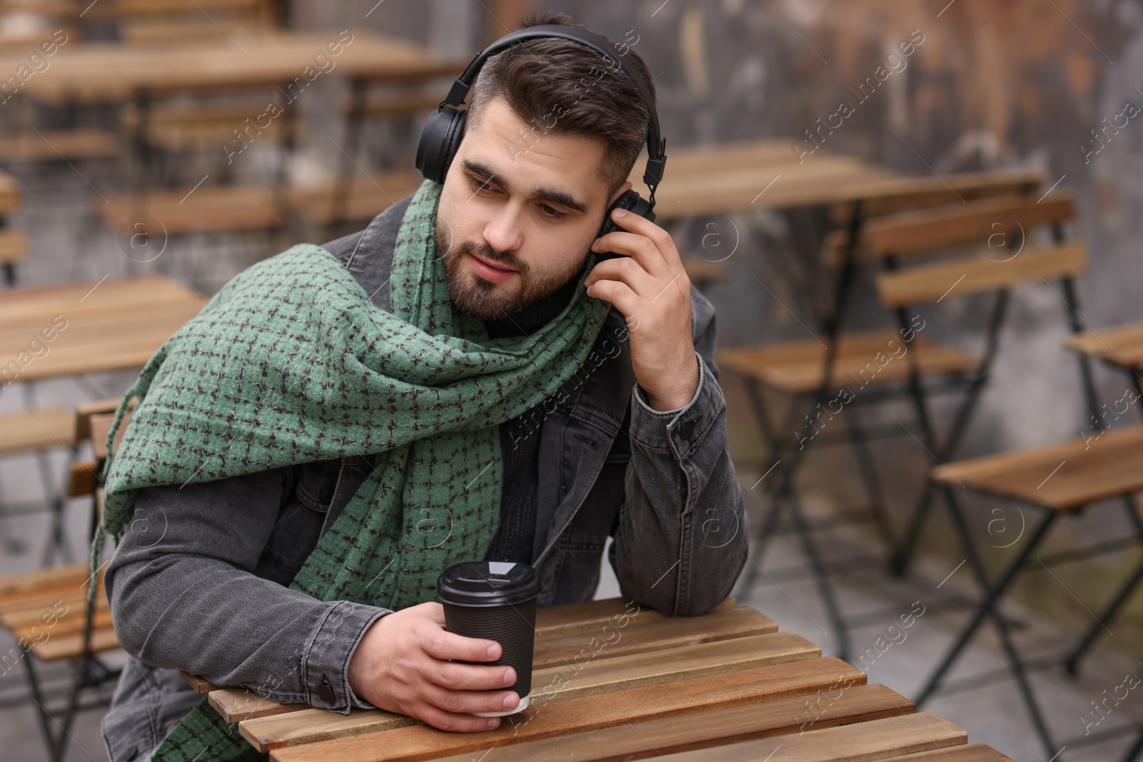 Photo of Handsome man in warm scarf with paper cup listening to music in outdoor cafe