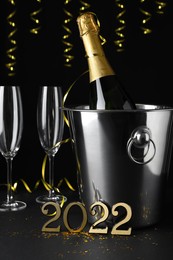 Photo of Happy New Year 2022! Bottle of sparkling wine in bucket and glasses on table against black background