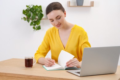 Photo of Happy young woman with notebook working on laptop at wooden table indoors