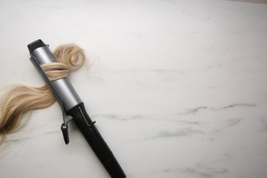 Curling iron with blonde hair lock on white marble background, top view. Space for text