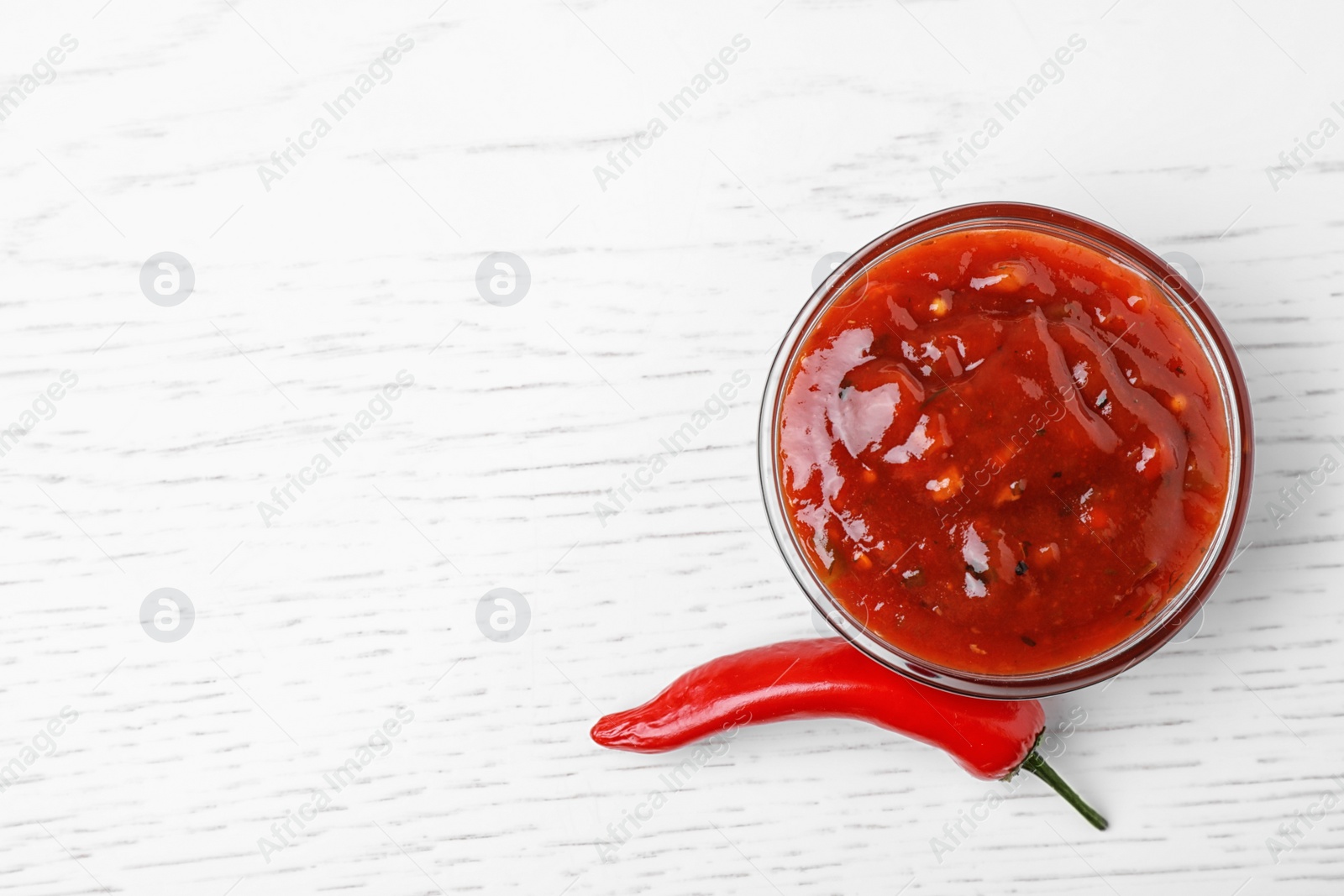 Photo of Bowl of hot chili sauce with red pepper on white wooden background, top view. Space for text