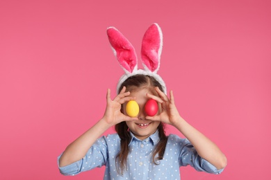 Photo of Little girl in bunny ears headband holding Easter eggs near eyes on color background