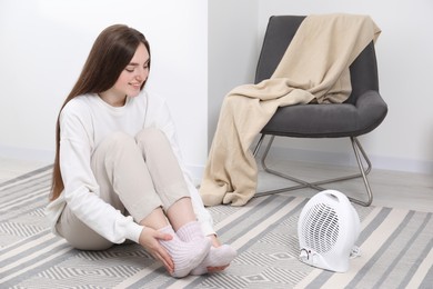 Young woman warming feet near electric fan heater at home