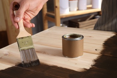 Man with brush and can applying wood stain onto wooden surface indoors, closeup