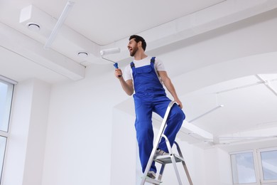 Photo of Handyman with roller having fun on step ladder in room, low angle view. Ceiling painting