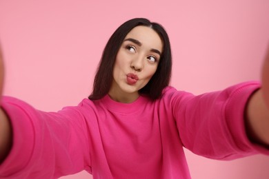 Young woman taking selfie and blowing kiss on pink background