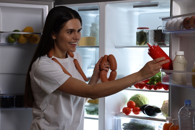 Photo of Young woman taking ketchup and sausages out of refrigerator at night