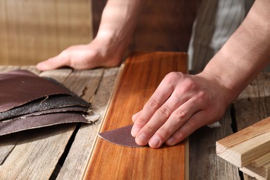 Photo of Man polishing wooden plank with sandpaper at table indoors, closeup