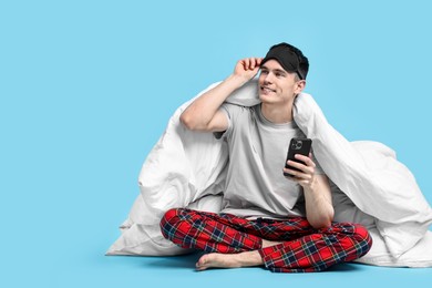 Happy man in pyjama wrapped in blanket using smartphone on light blue background, space for text