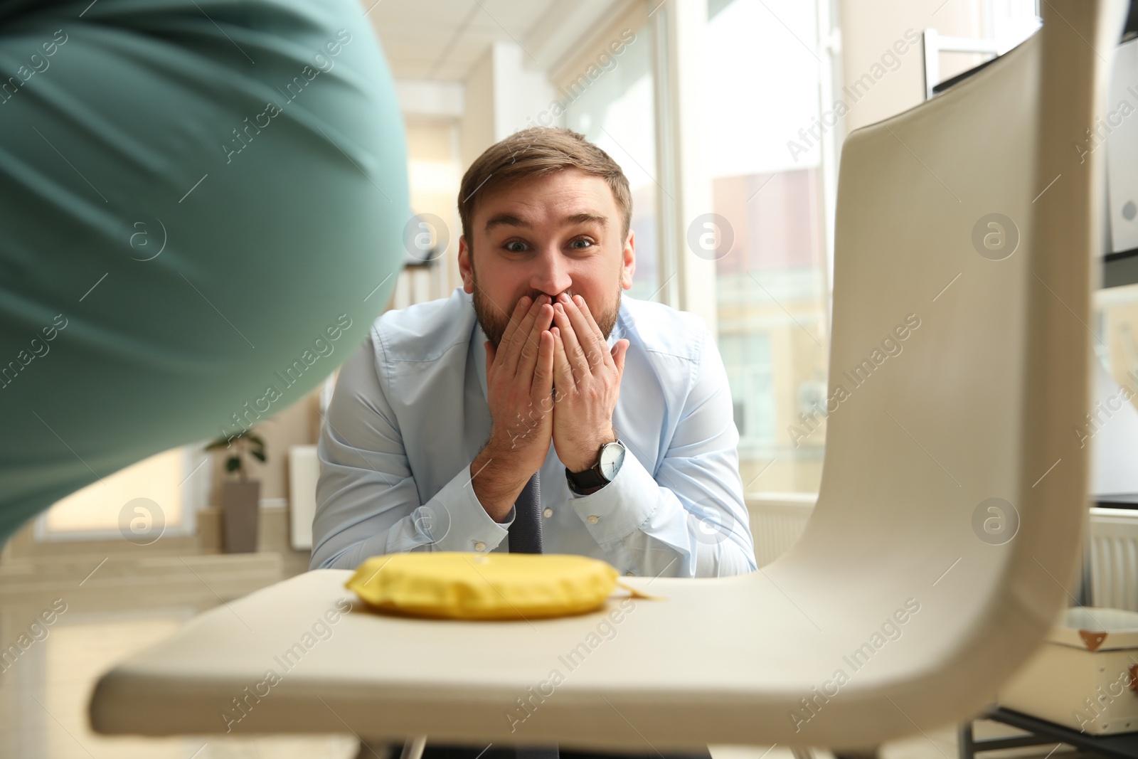 Photo of Young man putting whoopee cushion on chair while his colleague sitting down in office, closeup. Funny joke