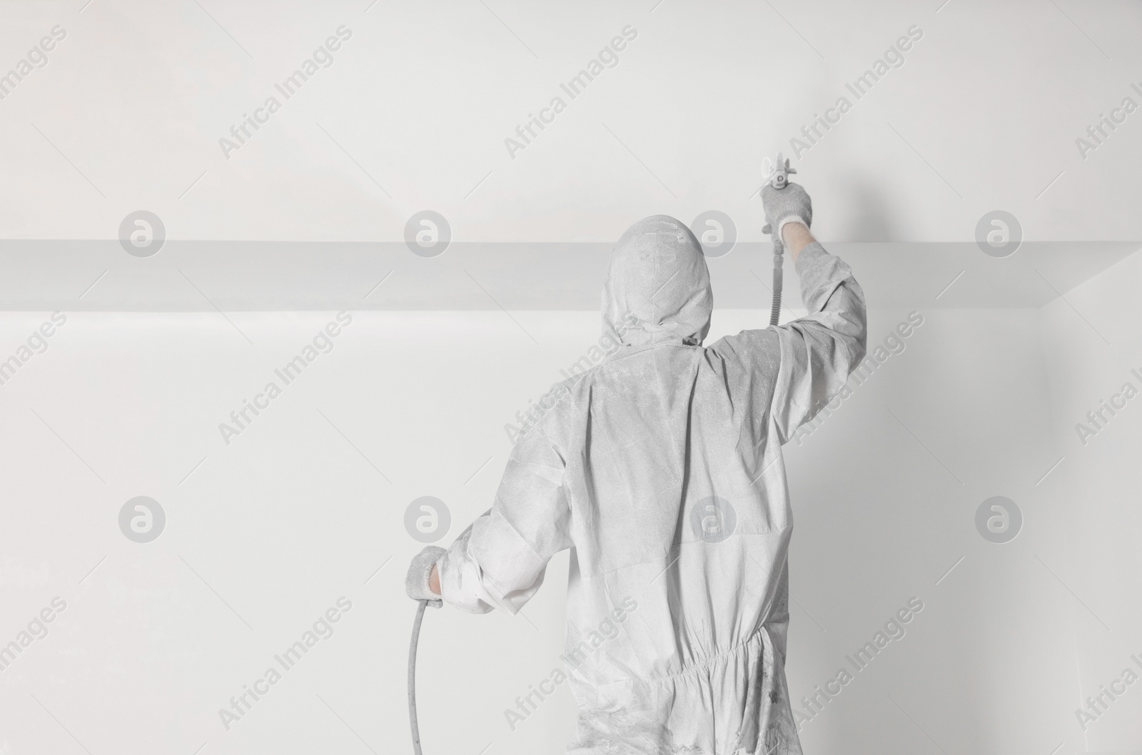 Photo of Decorator in protective overalls painting wall with spray gun indoors