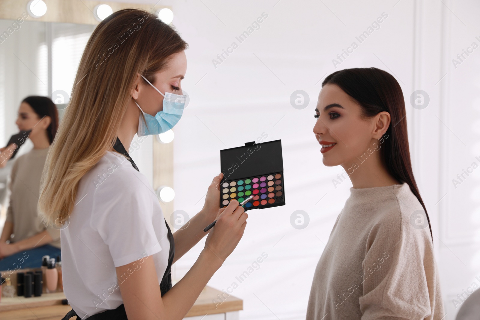 Photo of Makeup artist in protective mask working with woman indoors. Preventive measures during COVID-19 pandemic