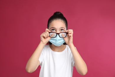 Photo of Little girl wiping foggy glasses caused by wearing medical face mask on pink background. Protective measure during coronavirus pandemic