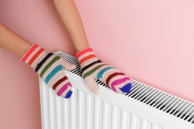 Photo of Woman in mittens warming hands on heating radiator near color wall