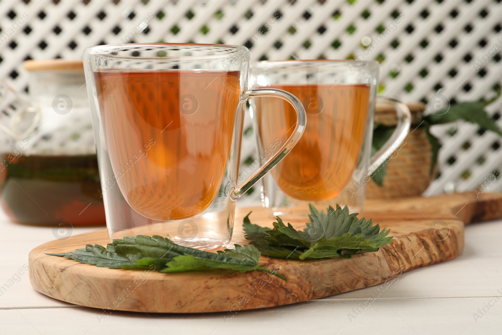 Photo of Glass cups of aromatic nettle tea and green leaves on white wooden table, closeup