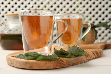 Glass cups of aromatic nettle tea and green leaves on white wooden table, closeup