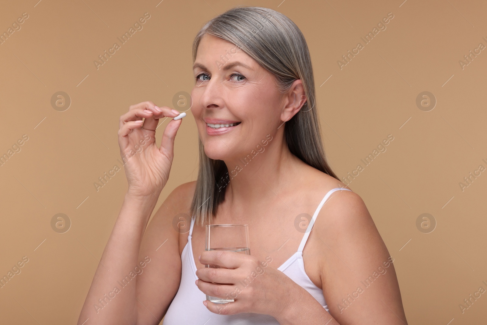 Photo of Beautiful woman with glass of water taking vitamin capsule on beige background