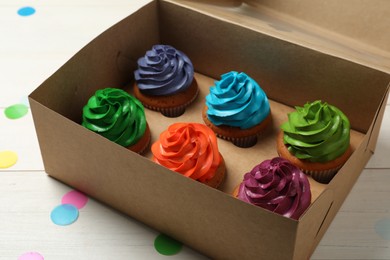 Photo of Box with different cupcakes and confetti on white wooden table, closeup
