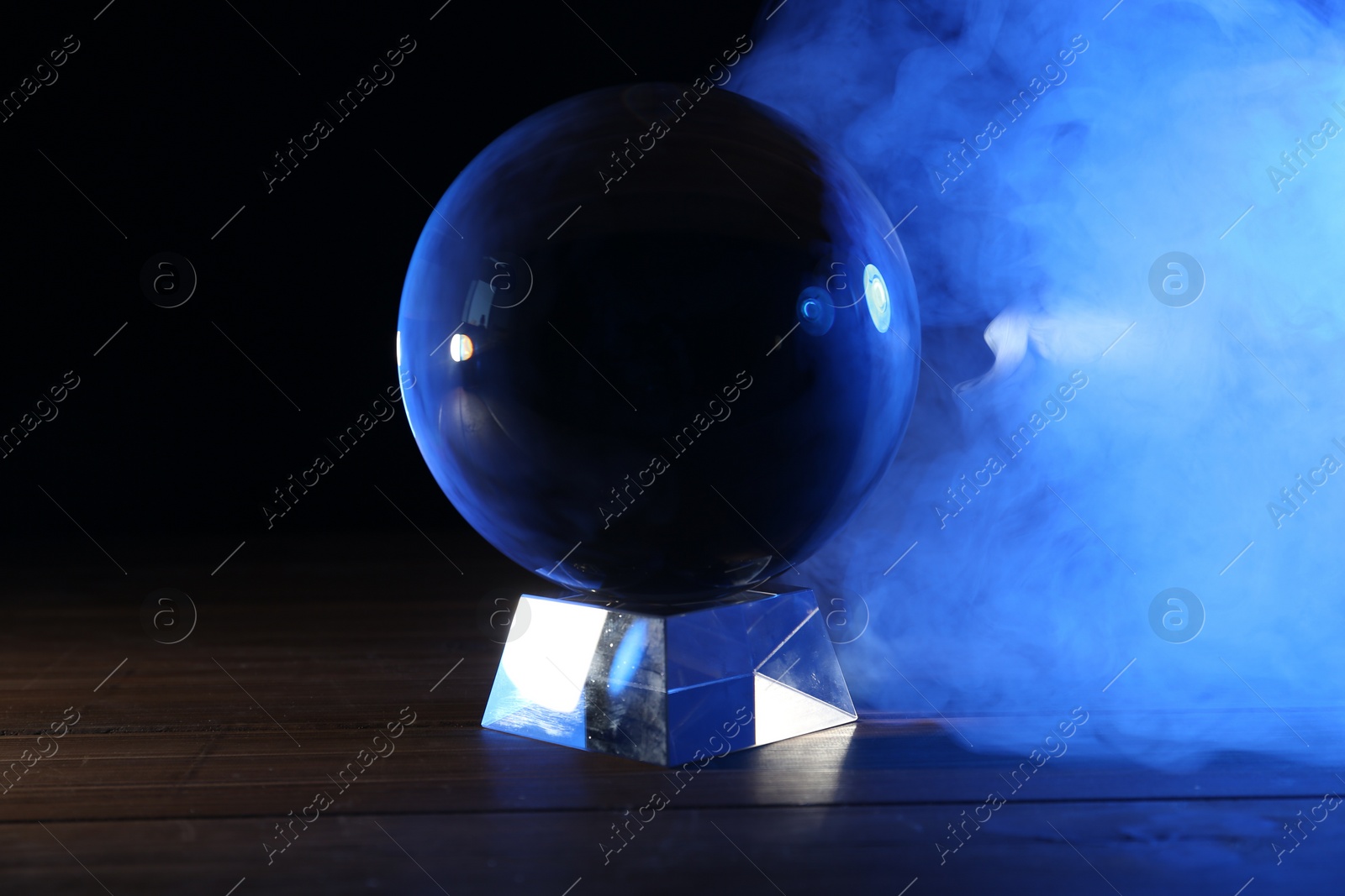 Photo of Magic crystal ball on wooden table and smoke against dark background. Making predictions