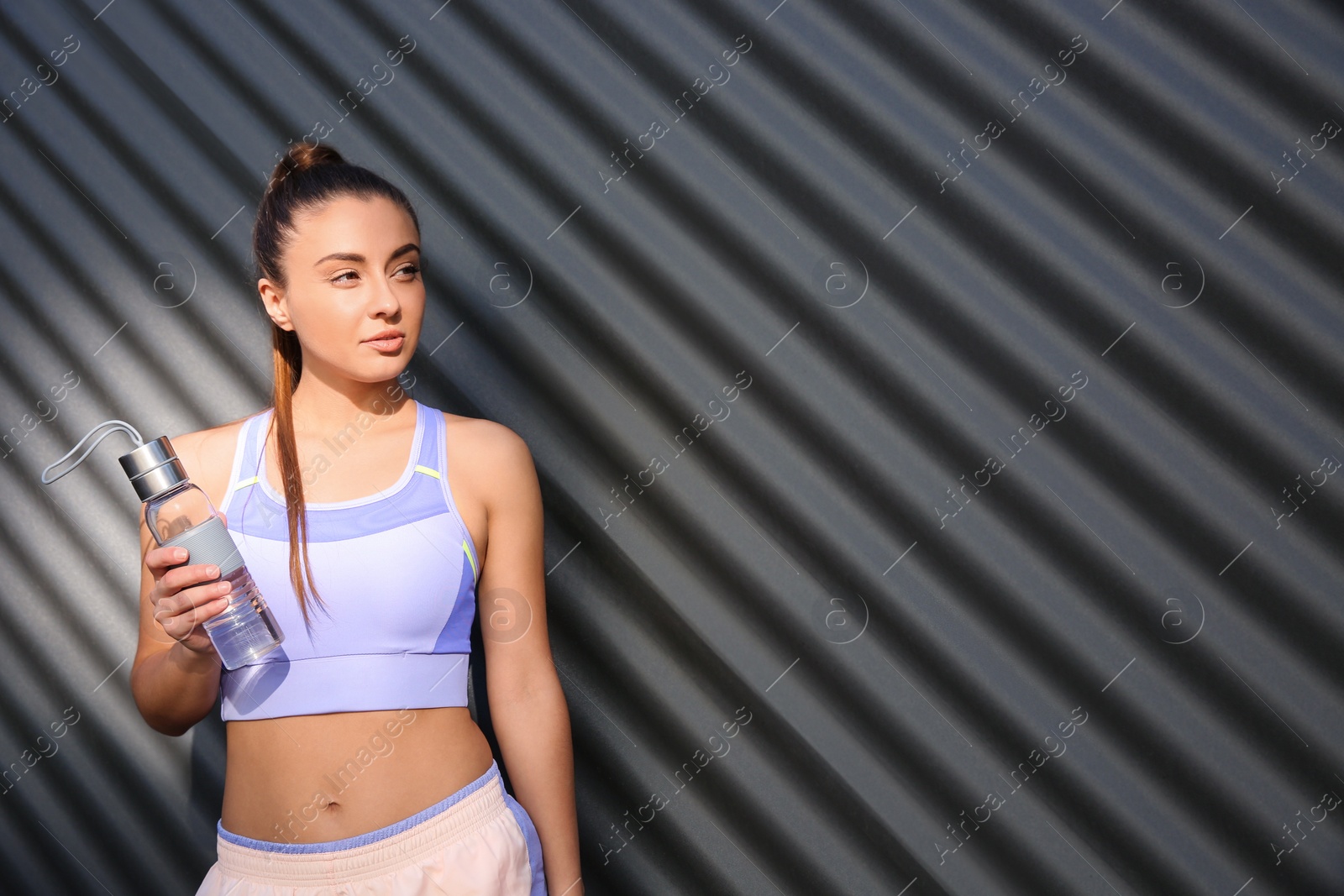 Photo of Young woman in sportswear with bottle near corrugated metal wall. Space for text