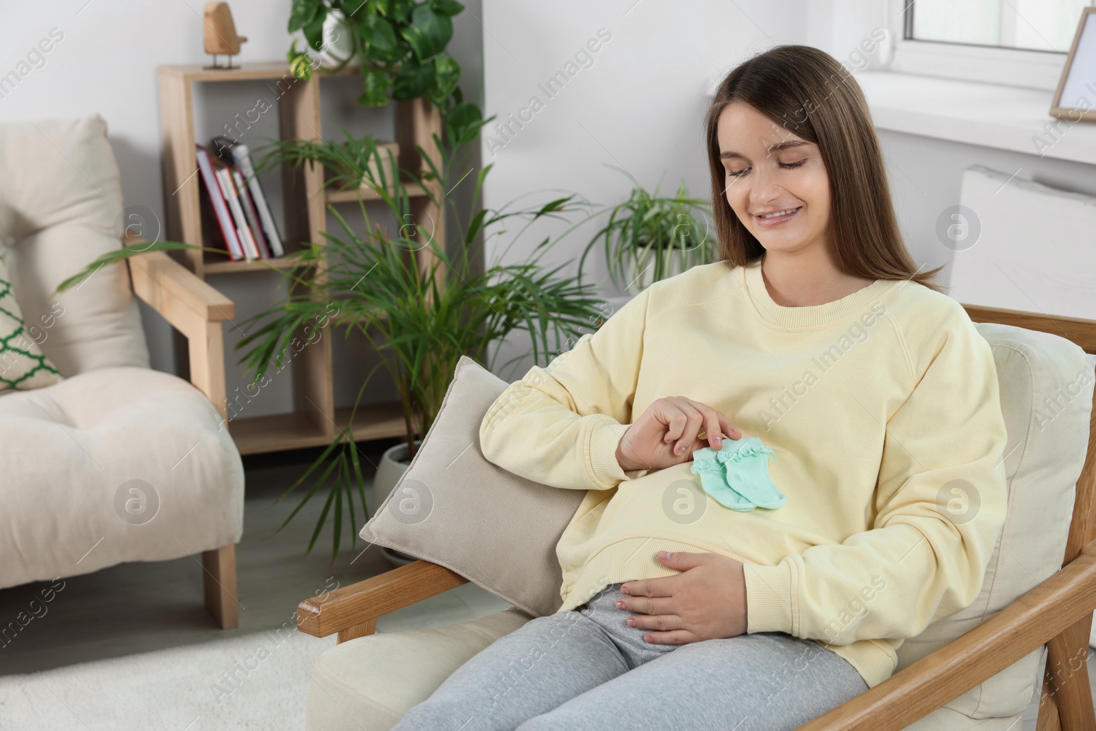 Photo of Happy pregnant woman sitting on armchair with baby socks in living room